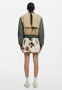 Mouwloze beige bomberjack met groene satijnen mouwen, gecombineerd met een witte rok met bloemenprint. Casual schoeisel en witte sokken zichtbaar.