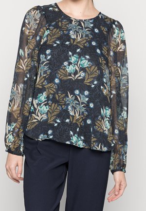 Die Bluse mit floralem Muster hat lange, durchsichtige Ärmel, einen runden Ausschnitt und einen dunklen Hintergrund mit blauen und grünen Blumenmustern. Sie wird mit dunklen Hosen kombiniert.