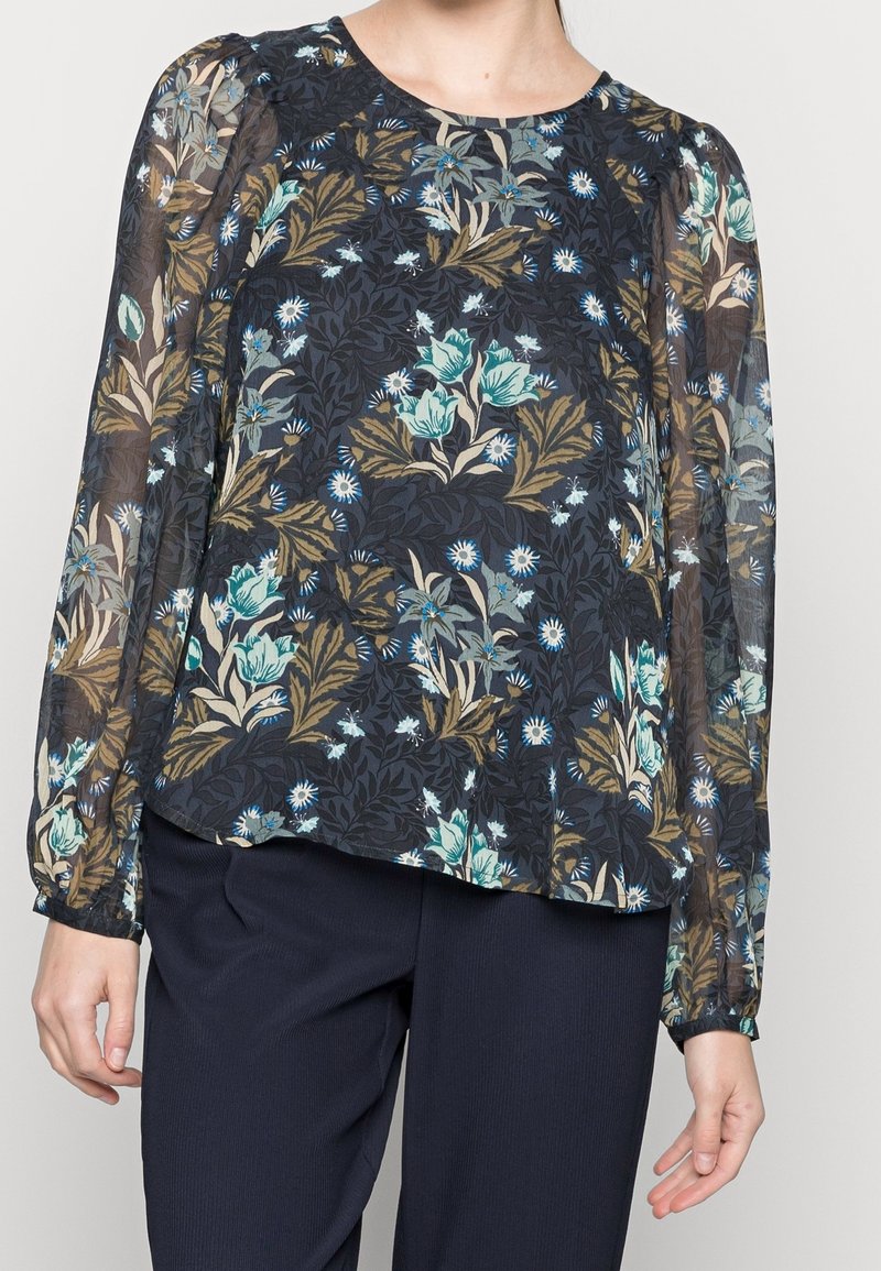 Blusa floreale con maniche lunghe trasparenti, scollatura rotonda e uno sfondo scuro decorato con motivi floreali blu e verdi. Abbinata a pantaloni scuri.