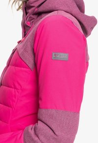 Giacca isolante rosa con parte superiore e maniche grigie melange. Presenta una toppa con il logo Roxy sul braccio e un colletto alto con cappuccio.