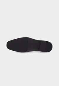 Suela de un zapato formal negro con patrones de agarre texturizados y un tacón ligeramente elevado sobre un fondo blanco simple.