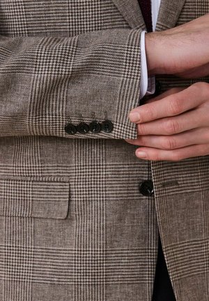 Mand justerer ærmet på brun ternet blazer med sorte knapper og hvid skjortemanchet synlig.