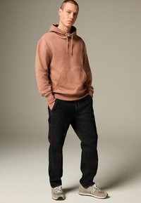 Hooded sweatshirt in dusty rose, relaxed fit, front pocket. Paired with black jeans, sleek cut, and light gray sneakers with dark accents.