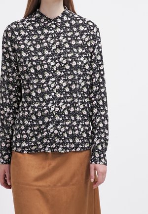 Femme portant un chemisier noir à fleurs à manches longues boutonné, associé à une jupe en daim marron, visible de la poitrine à mi-cuisse.