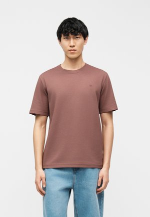 Junger Mann mit dunklem Haar, der ein schlichtes braunes T-Shirt mit kurzen Ärmeln und hellblaue Jeans trägt, vor einem schlichten weißen Hintergrund steht.