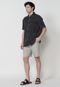 Camisa negra de manga corta con tejido texturizado, con cierre de botones en la parte delantera y bolsillo en el pecho, combinada con pantalones cortos gris claro y sandalias negras tipo slide.