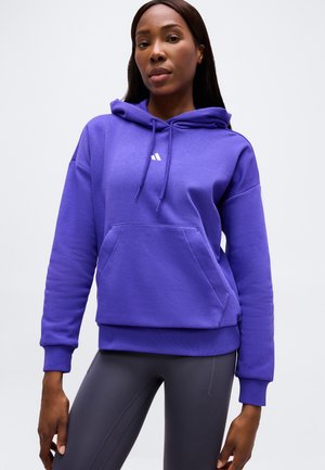 Femme portant un sweat à capuche violet avec des cordons et une poche avant, associée à des leggings gris foncé, debout devant un fond uni.