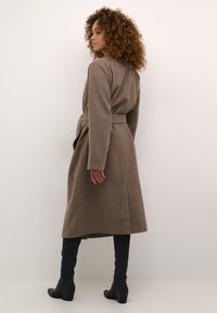 Brūns vilnas maisījuma mētelis ar garām piedurknēm, jostu vidukļa daļā, sānu kabatām un ceļa garuma apakšdaļu, valkāts virs tumšiem džinsiem un melnām kurpēm ar papēdi.