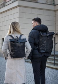 Two black leather backpacks displayed on individuals, featuring roll-top closures, adjustable straps, and metal buckles. Textured material.