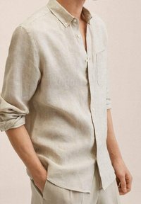 Chemise en lin beige clair à manches longues, col boutonné et poche poitrine unique. Texture douce avec une coupe décontractée et manches retroussées.