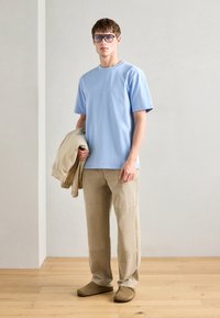 Ανοιχτό μπλε βαμβακερό t-shirt, μπεζ παντελόνι σε χαλαρή γραμμή, μπεζ bomber jacket κρεμασμένο από τ�ο χέρι, καφέ παντόφλες, γυαλιά με διαφανή σκελετό.