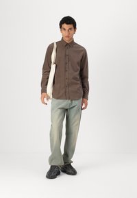 Braunes Button-Up-Hemd, hellblaue Jeans und schwarze Sneakers mit Muster. Das Model hält eine beigen Tragetasche und steht vor einem schlichten Hintergrund.