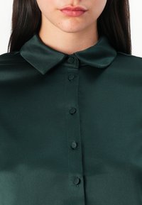 Blusa di satin verde scuro con colletto classico e chiusura frontale con bottoni. Texture liscia con bottoni rotondi distribuiti uniformemente.