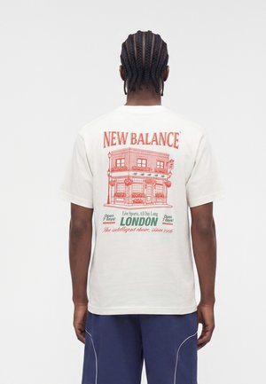 Persona con cabello trenzado que lleva una camiseta blanca de New Balance con gráfico de una tienda de Londres y texto en la espalda.
