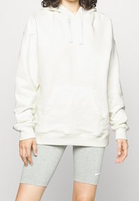 Sudadera con capucha blanca de algodón con un bolsillo frontal y cordón, combinada con pantalones cortos de ciclismo grises. La tela es suave y ligeramente texturizada.
