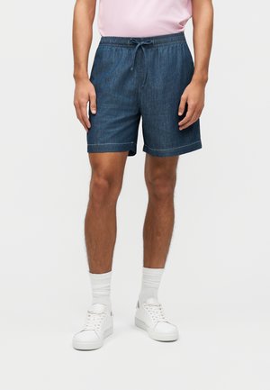 Homme portant un short à cordon de serrage bleu foncé, des chaussettes blanches, des baskets blanches et une chemise rose, debout devant un fond clair uni.