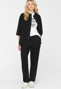 Young woman with blonde hair wearing a black buttoned shirt, black pants, white graphic t-shirt, and white sneakers, standing with one hand in pocket.
