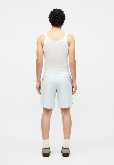 Άνδρας με λευκή φανέλα, ανοιχτό μπλε σορτς, λευκές κάλτσες και καφέ παπούτσια, όρθιος με την πλάτη προς ένα απλό λευκό φόντο.