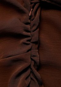 Hnědá tkanina s vertikálním řasením a bočním zipem. Textura vypadá hladce s jemným leskem, který zvýrazňuje nabíraný detail.