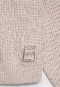 Beige gestrickter Stoff mit weicher Textur; verfügt über ein braunes Etikett mit dem Text "KNITTERS 99 SINCE 1991" und dekorativen Akzenten.