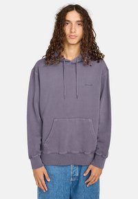Lila Baumwollkapuzenpullover mit einer Kängurutasche, Zugbändern und gerippten Bündchen. Besitzt ein dezentes Logo auf der Brust. Getragen mit blauen Jeans.