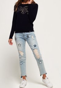 Laivastonsininen neulepaita, jossa metallinen "Superdry"-logo, vaaleansiniset kulutetut farkut tähtikuvioin ja valkoiset tennarit. Rentoon tyyliin sopiva asukokonaisuus, jossa monipuolisia tekstuureja.