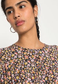 Blusa floral com fundo preto; apresenta pequenas flores multicoloridas, decote franzido e tecido leve. Brincos circulares prateados.