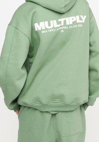 Persona che indossa un felpa verde chiaro e pantaloni abbinati con il testo "MULTIPLY MULTIPLY APPAREL CLUB 2024" in bianco sulla parte posteriore della felpa.
