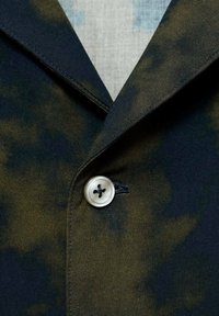 Marineblauwe en olijfgroen gepatteerde blazer met een geknipte kraag, enkele knoopsluiting en textuurstof. Beschikt over een ronde knoop met vier gaten.