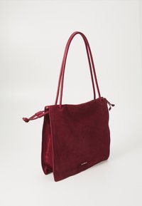 Sac à main en daim bordeaux avec deux longues anses assorties. Présente un design simple avec un fond plat et un branding minimaliste à l'avant.