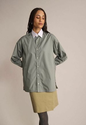 Femme aux cheveux tressés portant une chemise grise boutonnée surdimensionnée, un col blanc, une jupe beige et des collants foncés, debout les mains dans le dos.