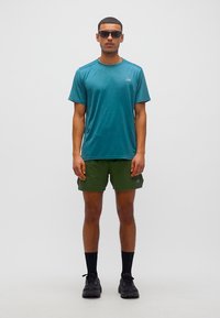 Άνδρας που φοράει πετρόλ αθλητικό μπλουζάκι, χακί σορτς, μαύρες κάλτσες και μαύρα αθλητικά παπούτσια, στέκεται μπροστά σε λευκό μονόχρωμο φόντο.