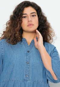 Vaaleansininen farkkupaita, jossa on kauluspanta, etunapit ja kerätty vyötärö. Omistaa pitkät hihat, joissa on halkiot hihansuissa.