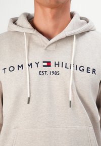 Man in een beige Tommy Hilfiger hoodie met een voorzak, witte trekkoorden en logo tekst "TOMMY HILFIGER EST. 1985" op de borst.