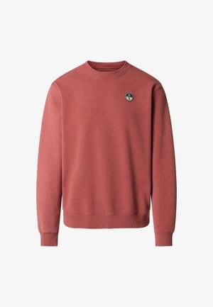 Homme portant des lunettes de soleil, un sweat-shirt rose poudré avec logo, un pantalon cargo crème et des baskets, debout avec une main dans la poche, regardant vers la gauche.