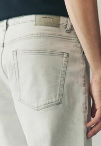 Svijetlosive traperice s klasičnim dizajnom stražnjeg džepa i kožnim logom označenim "DENIM SUPPLY" na pojasu.