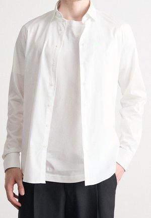 Homme portant une chemise blanche boutonnée par-dessus un t-shirt blanc et un pantalon noir, debout avec une main dans la poche sur un fond uni.