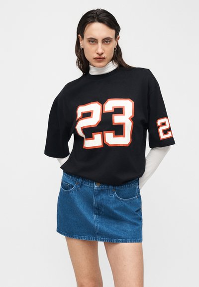 Νεαρή γυναίκα που φοράει μαύρο φαρδύ t-shirt με λευκή και κόκκινη εκτύπωση "23", λευκό μακρυμάνικο τοπ από μέσα και μίνι φούστα από μπλε τζιν.