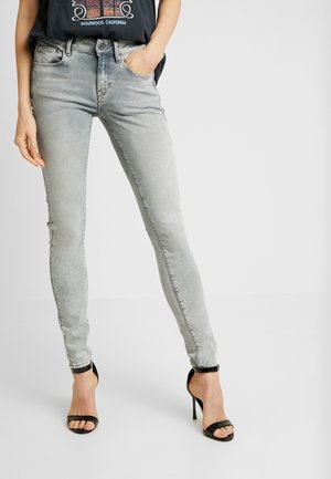 Jeans ajustados en gris desvanecido con una cintura de altura media, que cuentan con un clásico diseño de cinco bolsillos y un sutil detalle de desgaste, combinados con tacones negros.