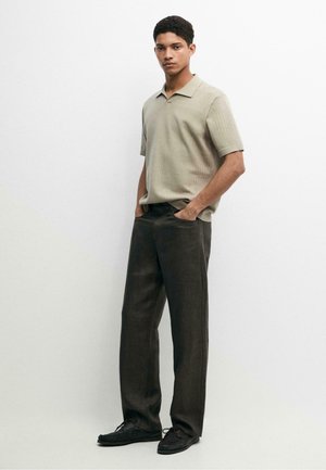 Jeune homme debout, les mains dans les poches, portant un polo en maille beige à manches courtes et un pantalon large marron foncé avec des chaussures noires.