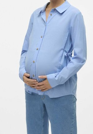 Gravid person iført en lys blå skjorte med knapper og blå jeans, hænderne blidt hvilende på maven mod en ensfarvet hvid baggrund.
