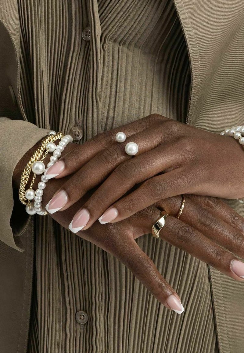 Pärlade armband med vita pärlor och guldaccenter, parat med avlånga, nudefärgade naglar med vita toppar, placerade på en strukturerad tygbakgrund.