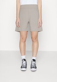 Beige Freizeithosen mit weitem elastischem Bund, zwei Seitentaschen und lockerem Schnitt, kombiniert mit schwarzen High-Top-Sneakern und weißen Socken.