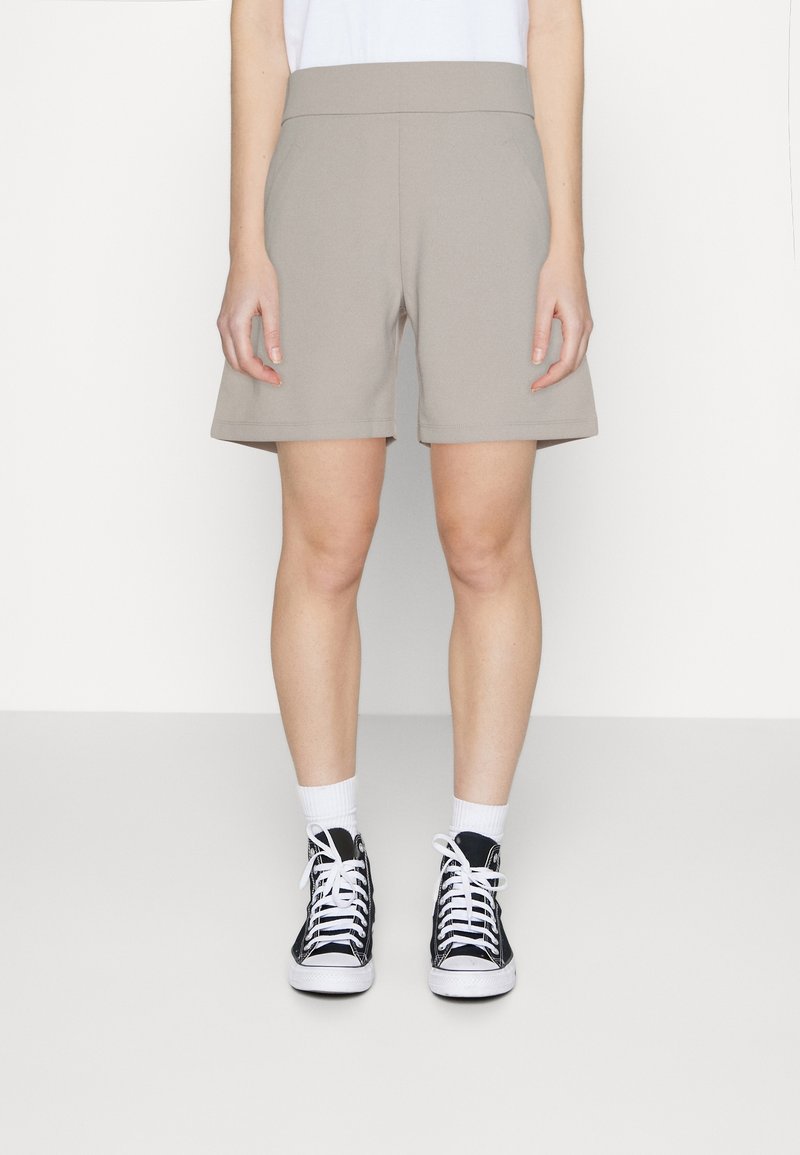 Beige Freizeithosen mit weitem elastischem Bund, zwei Seitentaschen und lockerem Schnitt, kombiniert mit schwarzen High-Top-Sneakern und weißen Socken.