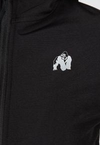 Melna kapucīna džemperis ar zīdainu tekstūru, ar izteiksmīgu sudraba gorillas logo kreisajā krūšu daļā.