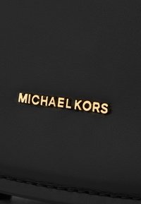 Materiale nero testurizzato con dettagli del logo "MICHAEL KORS" in oro. La finitura liscia e gli accenti di cucitura esaltano il design.