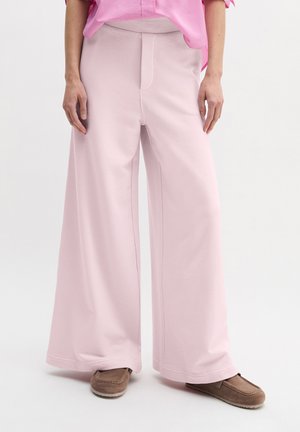 Osoba ubrana w jasnoróżowe spodnie o szerokich nogawkach, różową koszulę i brązowe wsuwane buty, stojąca na tle jednolitego białego tła.