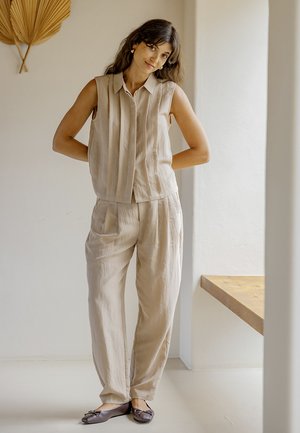 Vrouw met lang donker haar die een beige mouwloos topje en bijpassende losse broek draagt, staand binnen bij een houten tafel en wanddecoratie.