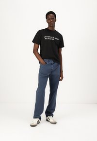 Svart bomulls t-skjorte med hvit tekst, kombinert med blå denim jeans. Modellen står foran en nøytral bakgrunn, iført hvite joggesko med svarte striper.