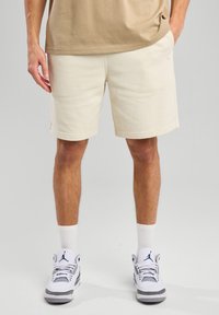 Beige shorts yang terbuat dari campuran katun dengan pinggang elastis dan saku samping. Dipadukan dengan sepatu sneakers putih dan abu-abu yang memiliki aksen hitam.